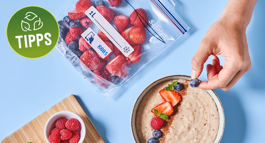Hand garniert eine Schale Porridge mit frischen Beeren. Im Hintergrund liegt ein Gefrierbeutel mit gefrorenen Beeren auf einem hellblauen Tisch. Links ist ein Holzbrett mit einer kleinen Schale Himbeeren zu sehen. 