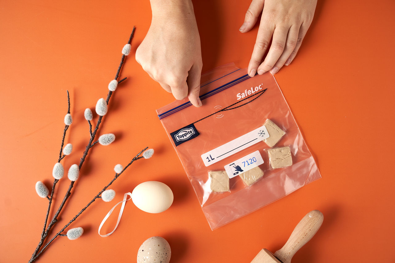 Zwei Hände verschließen einen Toppits SafeLoc Beutel mit mehreren Stücken frischer Hefe auf einem orangefarbenen Untergrund. Daneben liegen ein Weidenzweig mit Kätzchen, ein Osterei und ein Nudelholz