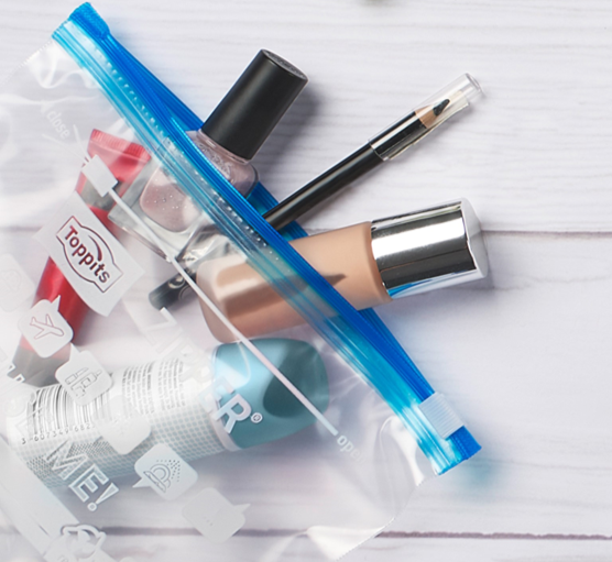 Transparenter Zipper-Beutel von Toppits, gefüllt mit Kosmetikartikeln wie Nagellack, Make-up und einem Pinsel, auf einem weißen Untergrund 